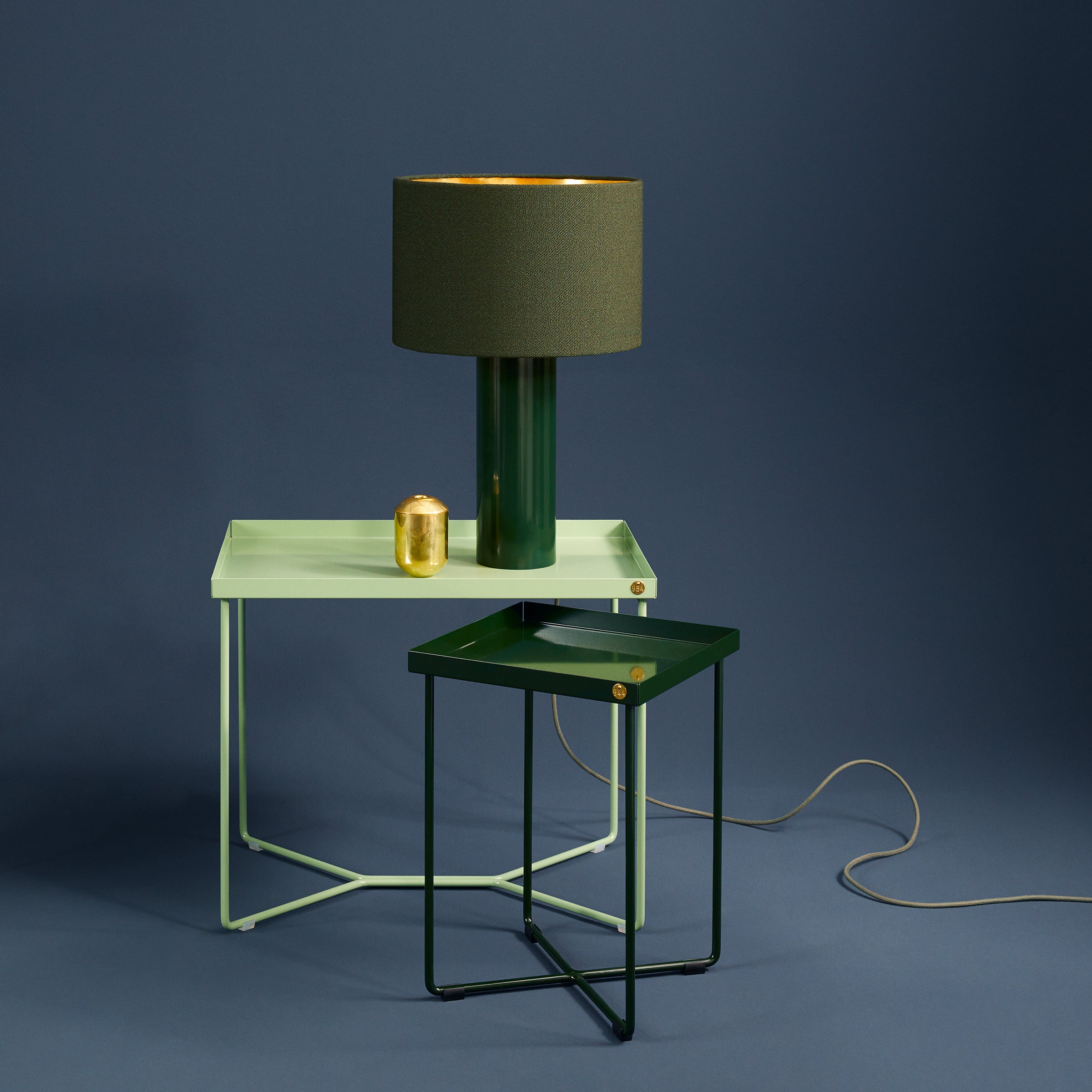 The Victor Foxtrot ALL ROUND Table Lamp pairs stylishly with two modern tables: one light green with a dark green lamp featuring a wool fabric lampshade, and the other table is dark green against a blue background. A small gold container enhances this sleek, minimalist design.