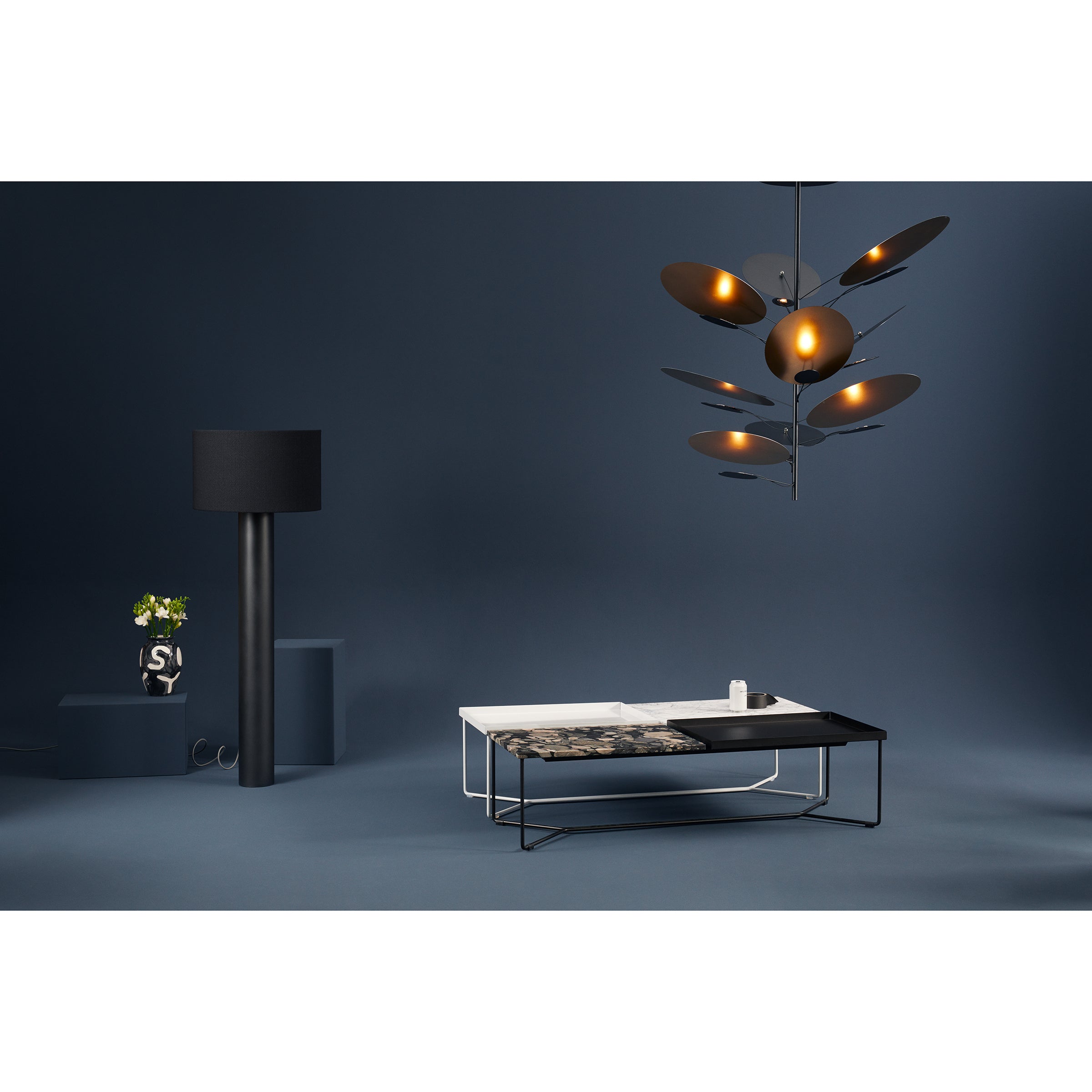 A modern living room showcases the ALL ROUND Floor Lamp by Victor Foxtrot, a geometric orb chandelier, and a minimalist coffee table. A small plant in a vase rests on a dark pedestal. The room glows warmly with a sleek, dark blue color scheme.