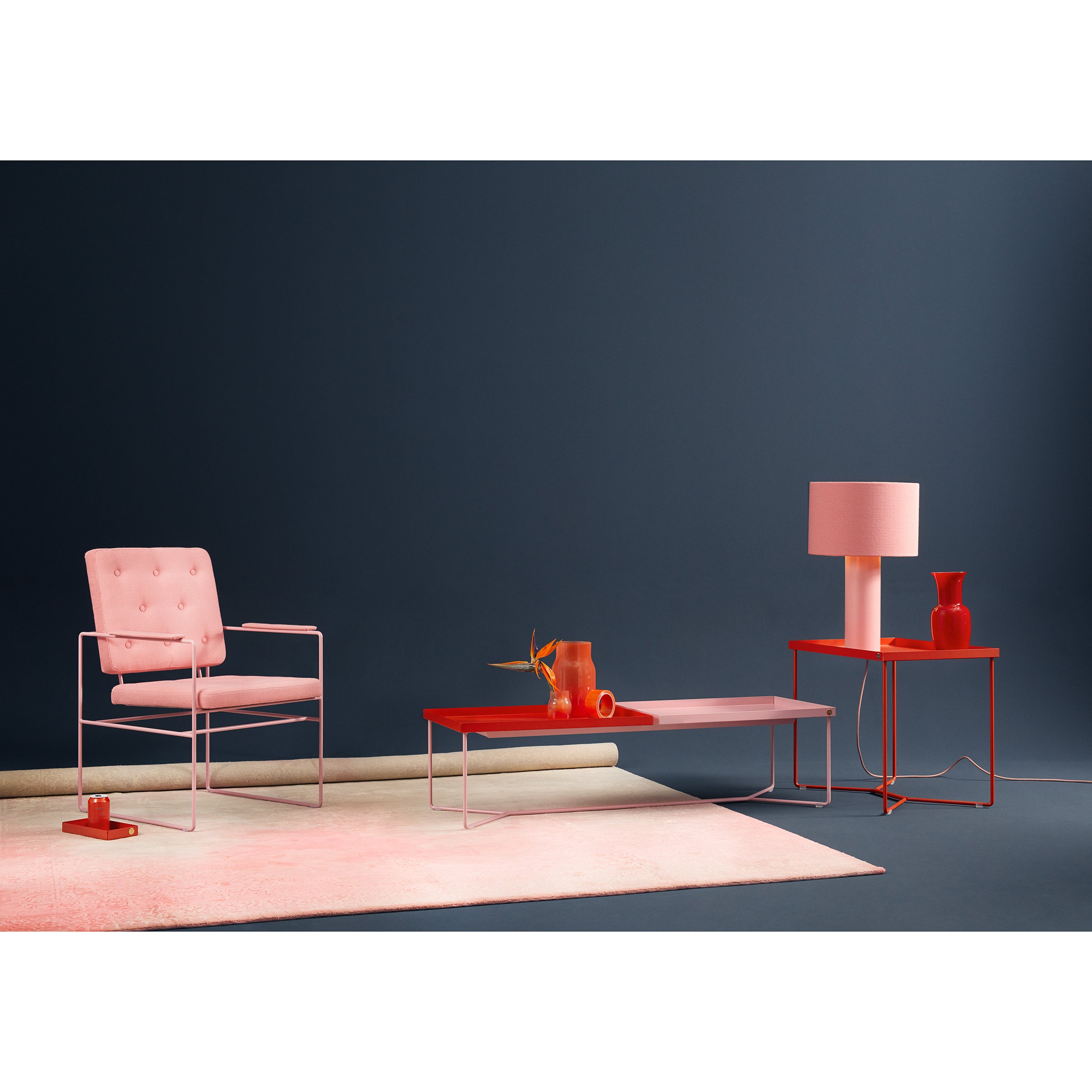 A modern living room with pink and red accents showcases a POI DOUBLE Coffee Table by Victor Foxtrot, featuring a sleek steel frame and leather inlays. It pairs well with a pink chair, red side table with a pink lamp, vases against the blue wall, and a soft pink rug on the floor.