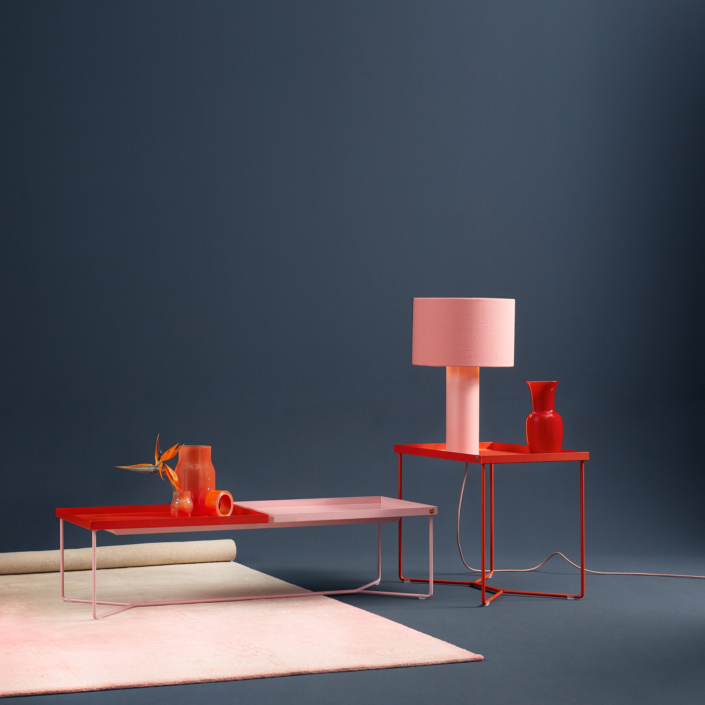 The Victor Foxtrot POI SINGLE Side Table, matching a modern red coffee table in powder-coated steel, stands against a dark blue backdrop. The coffee table displays red decorations while the side table holds a pink lamp and red vase, with a rolled-up rug nearby completing the setup.