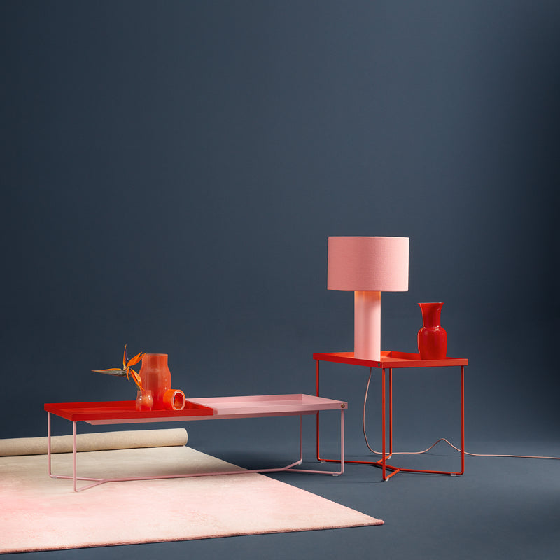 The Victor Foxtrot POI SINGLE Side Table, matching a modern red coffee table in powder-coated steel, stands against a dark blue backdrop. The coffee table displays red decorations while the side table holds a pink lamp and red vase, with a rolled-up rug nearby completing the setup.