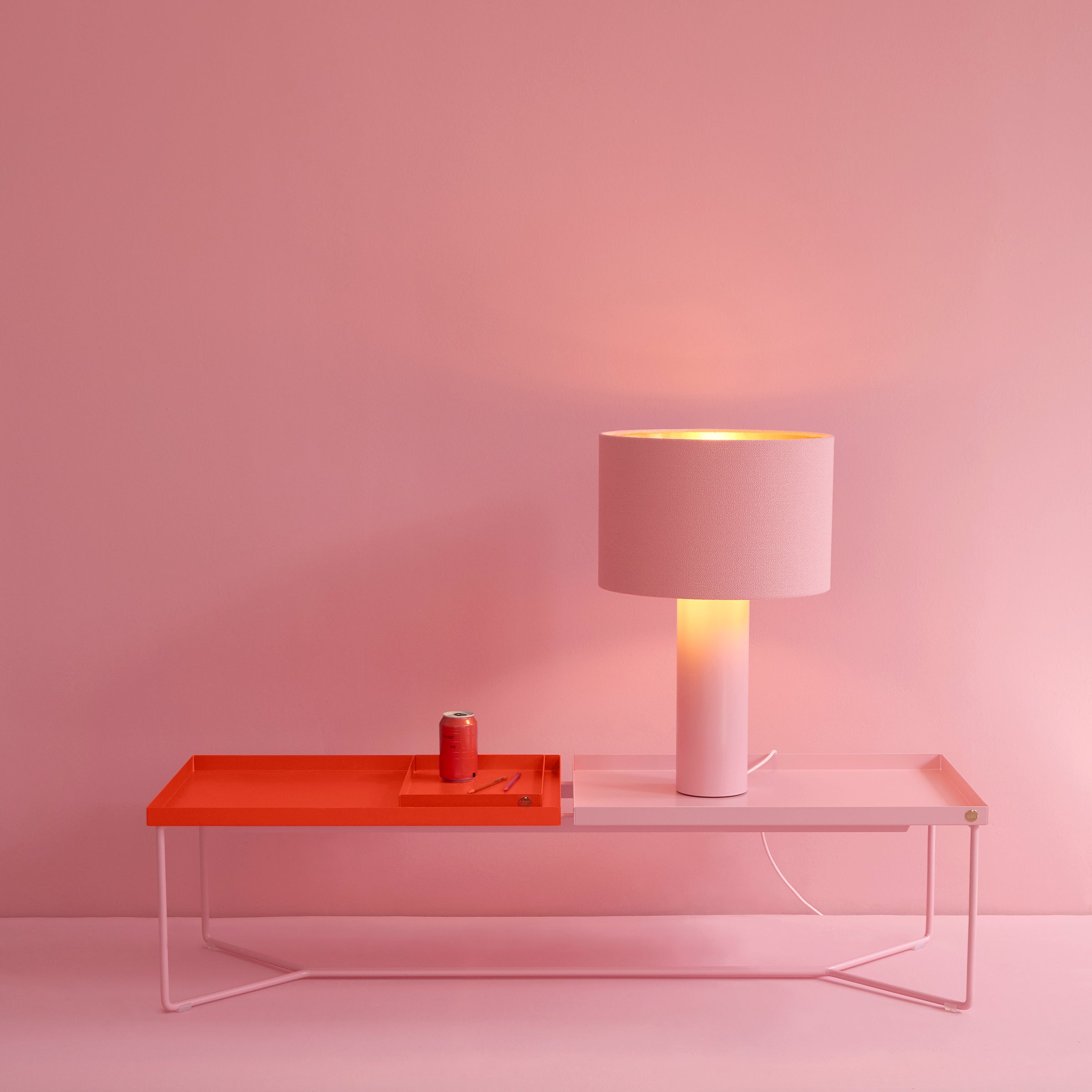 In a minimalist setting with a pink backdrop, the Victor Foxtrot ALL ROUND Table Lamp, featuring a wool fabric lampshade and powder-coated steel base, rests on a pink and orange table beside a small pink can. The scene is bathed in soft pink and orange tones.