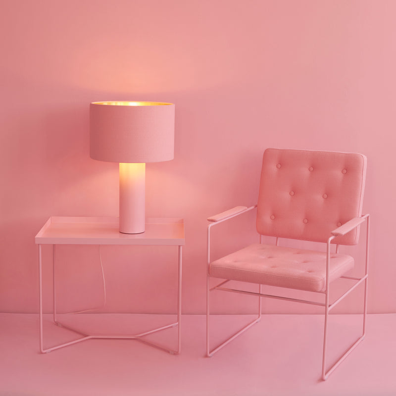 A pink room with a modern, minimalist design features a Victor Foxtrot POI SINGLE Side Table made of powder-coated steel, along with a pink table lamp and cushioned chair. The pink walls enhance the monochromatic theme, crafting a soft and cohesive look.