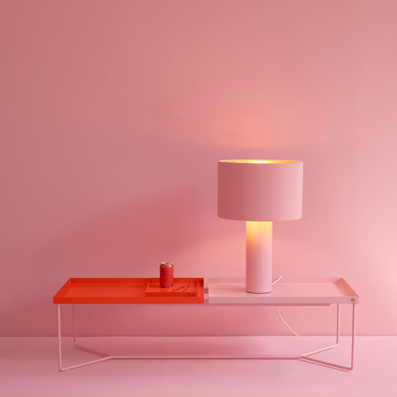 In a minimalist setting, a Victor Foxtrot POI DOUBLE Coffee Table with a steel frame is set against a pink wall. It holds a pink lamp casting warm light and a small red can on a tray, beautifully highlighting the scenes pink and red hues.