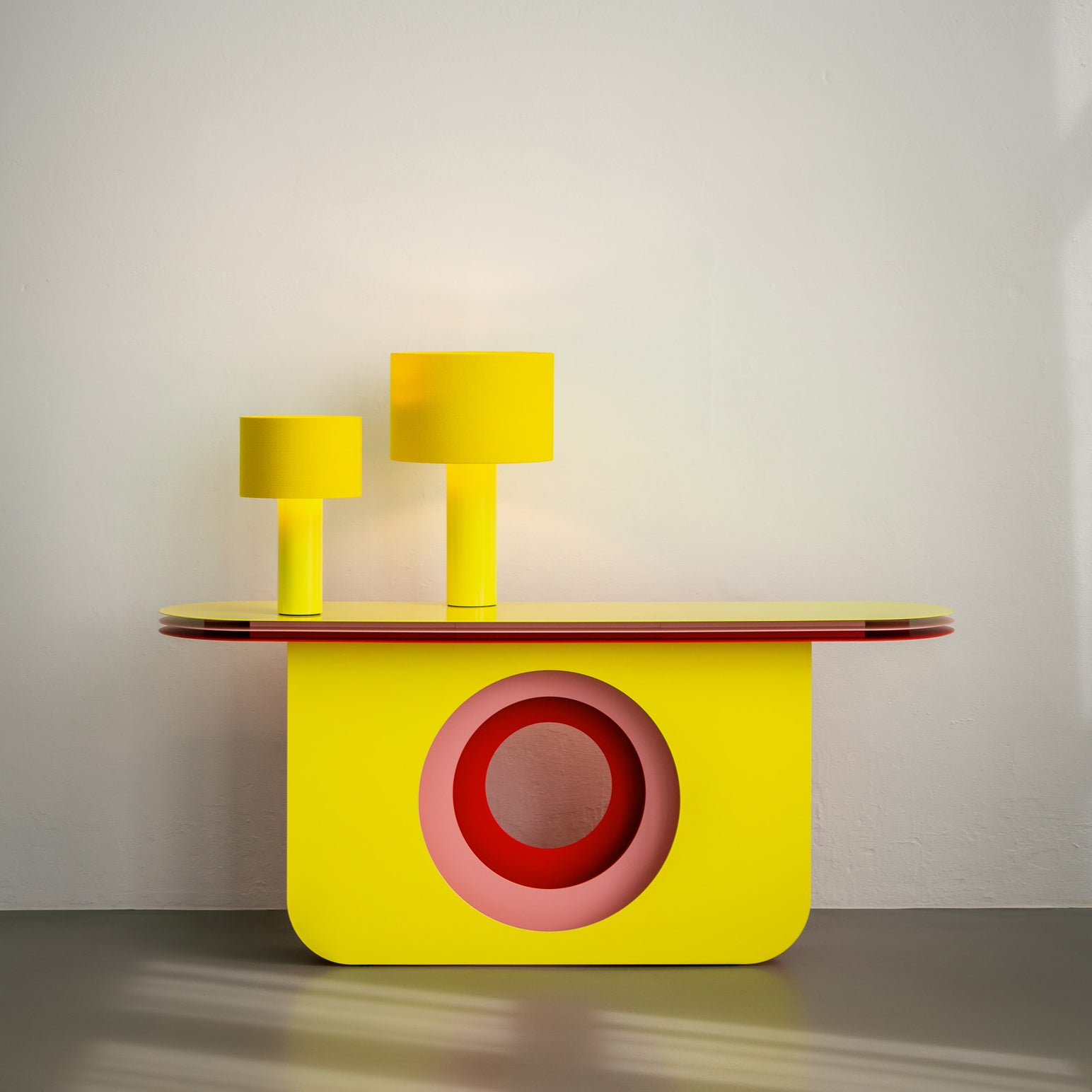 A sleek, artistic side table features a vibrant yellow finish and striking red circular accent. Topped with Victor Foxtrots ALL ROUND MINI Table Lamps in different heights, it elegantly contrasts the simple light gray walls and floor.