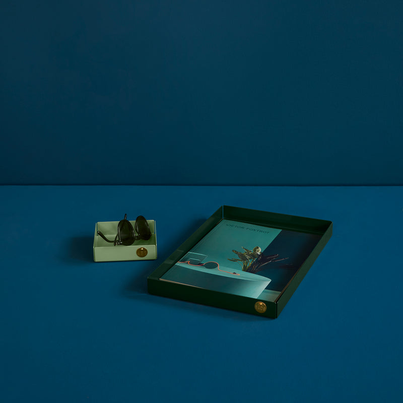 A dark green POI Tray L by Victor Foxtrot holds a botanical print book and teal envelope. Beside it, a lighter green steel box contains dark sunglasses. Set against a deep blue background, this minimalist arrangement reflects the brands design ethos.