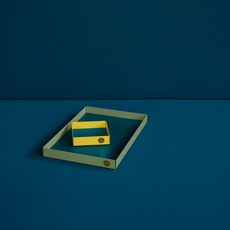 A yellow powder-coated steel box with a blue interior rests in a larger green POI Tray L by Victor Foxtrot. Set against a dark teal backdrop, they create a minimalist and colorful geometric composition, both adorned with the Victor Foxtrot emblem.