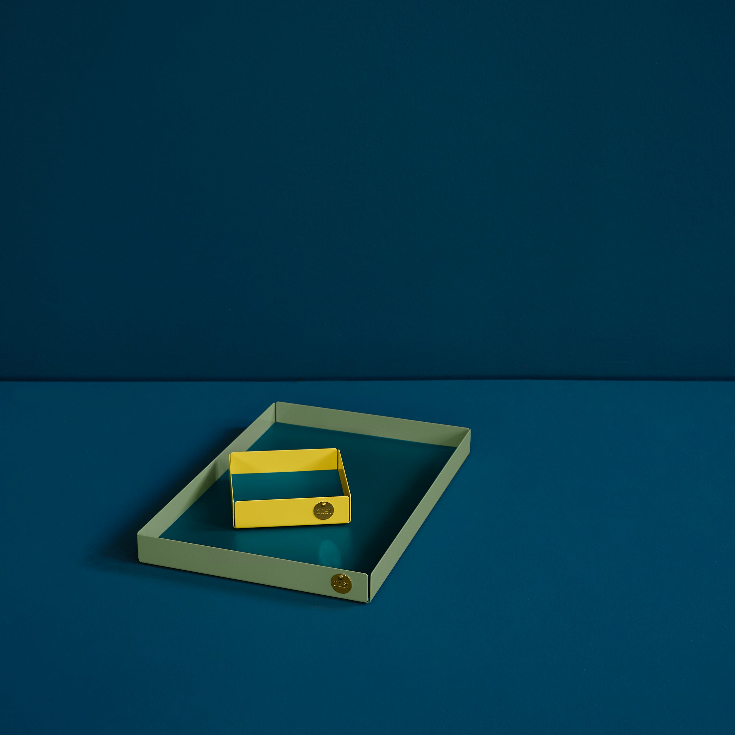Two rectangular POI trays from Victor Foxtrot rest on a dark teal surface. Design lovers will appreciate the olive green tray cradling its smaller yellow counterpart, both made from powder-coated steel with a round gold emblem. The minimalist scene highlights shape and color.