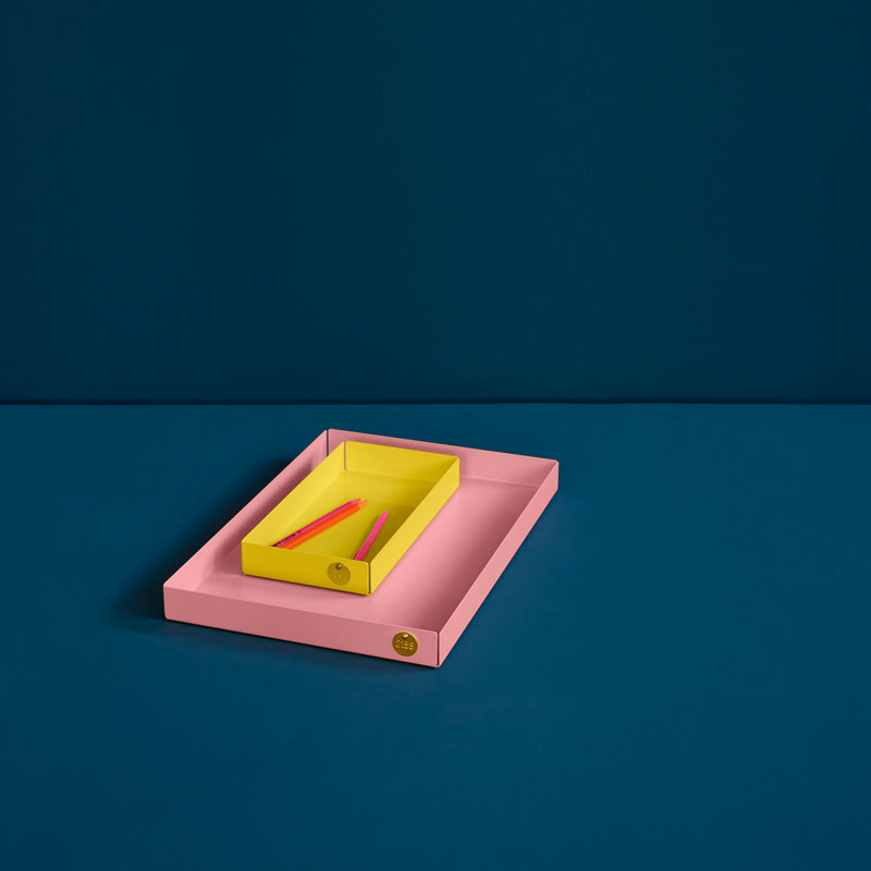 A minimalistic pink POI Tray L by Victor Foxtrot, crafted from powder-coated steel, rests on a blue surface. The smaller yellow tray nests inside, holding two red pens against a deeper blue backdrop that enhances its sleek design.