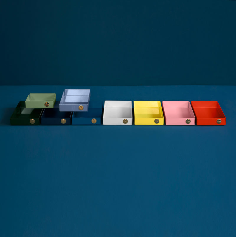 Eight colorful, open-top POI Tray M pieces by Victor Foxtrot, made from powder-coated steel in dark green, dark blue, light blue, white, yellow, pink, red, and orange, are displayed on a blue surface. Perfect for design enthusiasts with their small emblem detail.