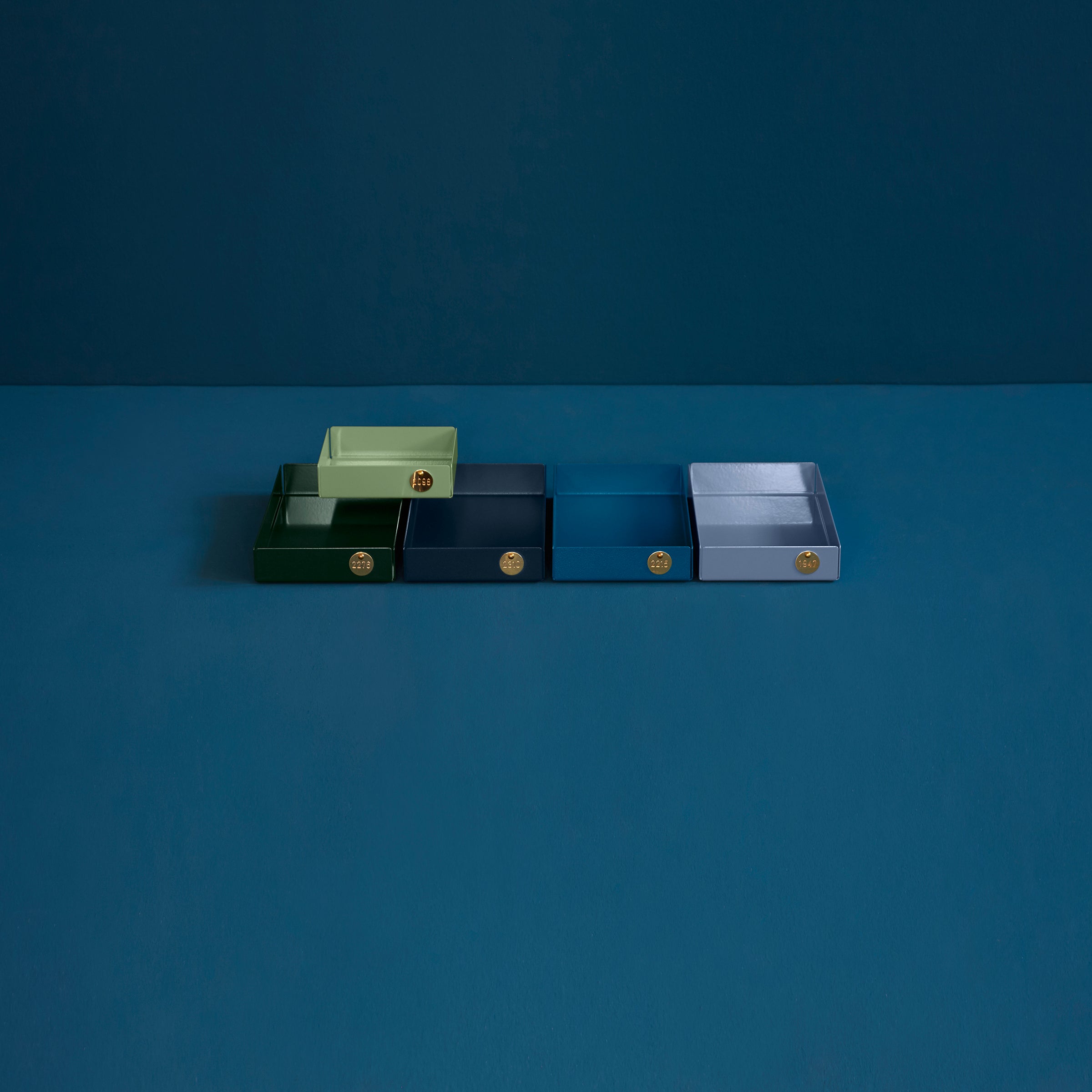 Design enthusiasts will admire Victor Foxtrots POI Tray M, featuring five blue and one green rectangular boxes on a dark surface. Crafted from powder-coated steel, each box has a round brass handle on a matching dark blue background.