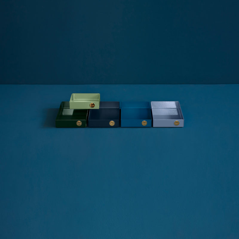 Design enthusiasts will admire Victor Foxtrots POI Tray M, featuring five blue and one green rectangular boxes on a dark surface. Crafted from powder-coated steel, each box has a round brass handle on a matching dark blue background.