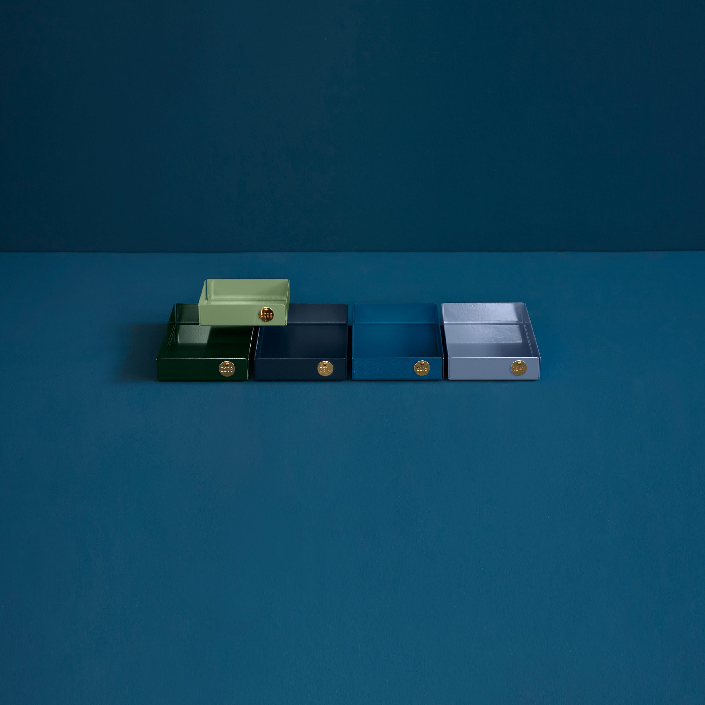 Five POI Tray M rectangular trays by Victor Foxtrot, crafted from powder-coated steel, are aligned on a blue surface for design lovers to admire. Each tray shines in pale green, dark green, blue, dark blue, and gray hues with a small gold emblem on the front—a tribute to modern elegance.