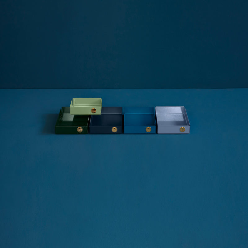 Five POI Tray M rectangular trays by Victor Foxtrot, crafted from powder-coated steel, are aligned on a blue surface for design lovers to admire. Each tray shines in pale green, dark green, blue, dark blue, and gray hues with a small gold emblem on the front—a tribute to modern elegance.