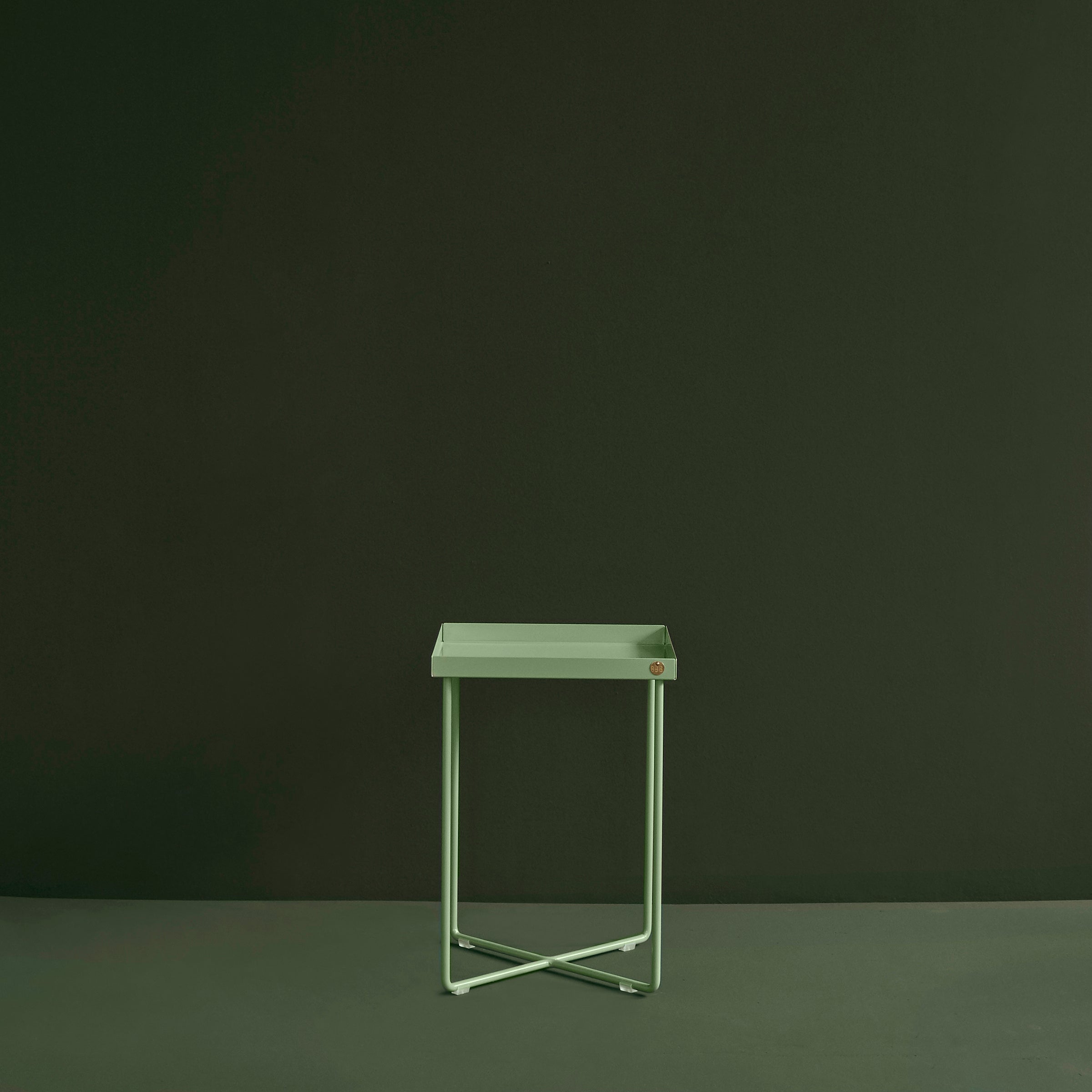The Victor Foxtrot POI MINI Side Table, featuring a minimalist light green square top with crisscross legs, stands out against a dark green backdrop.