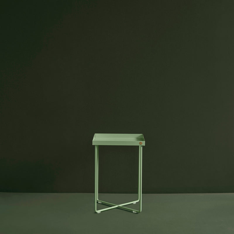 The Victor Foxtrot POI MINI Side Table, featuring a minimalist light green square top with crisscross legs, stands out against a dark green backdrop.