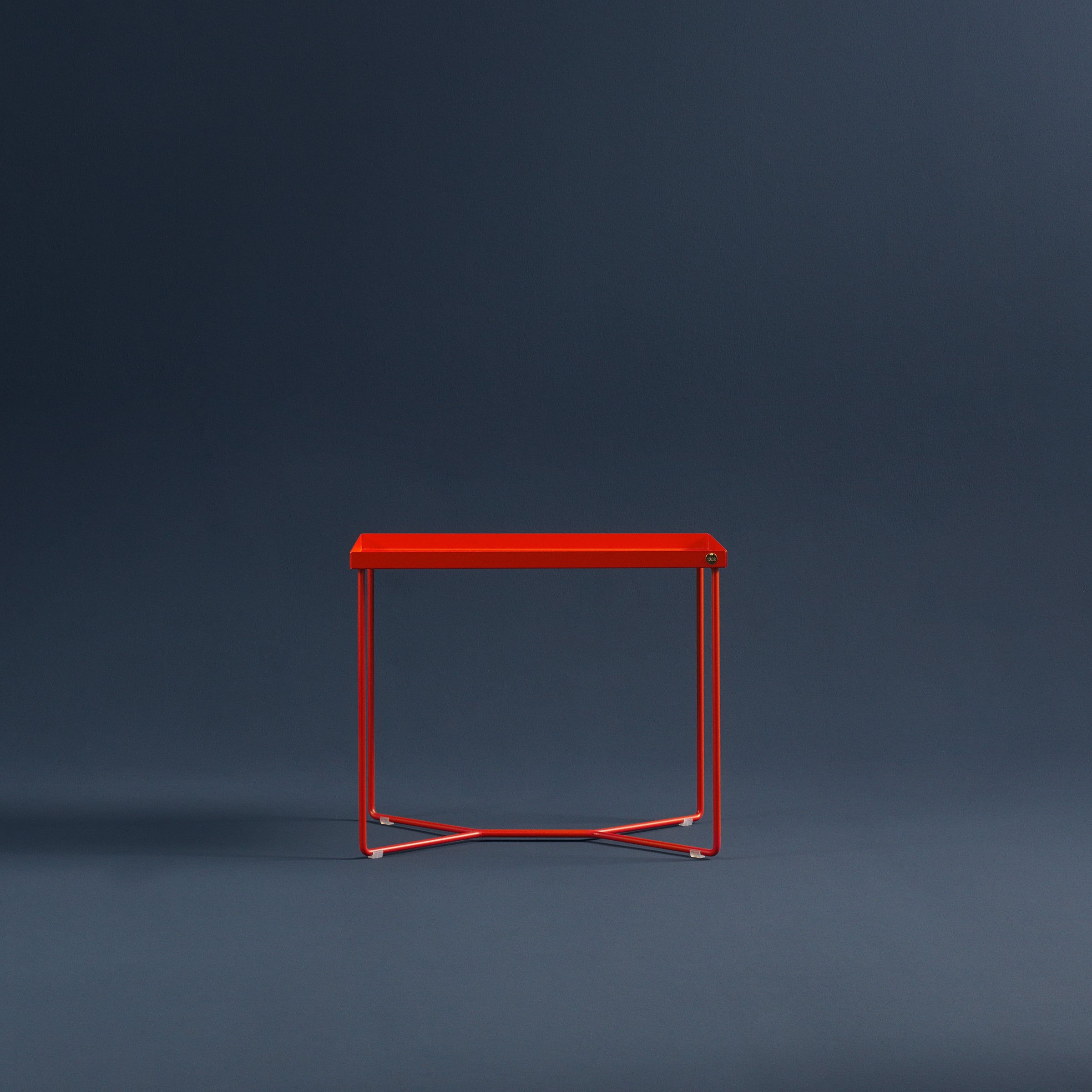 The POI SINGLE Side Table by Victor Foxtrot is a minimalist red piece made of powder-coated steel and slim metal legs, showcased against a dark blue backdrop.