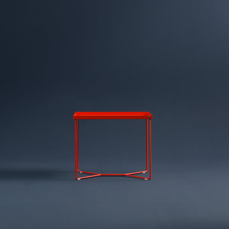 The POI SINGLE Side Table by Victor Foxtrot is a minimalist red piece made of powder-coated steel and slim metal legs, showcased against a dark blue backdrop.