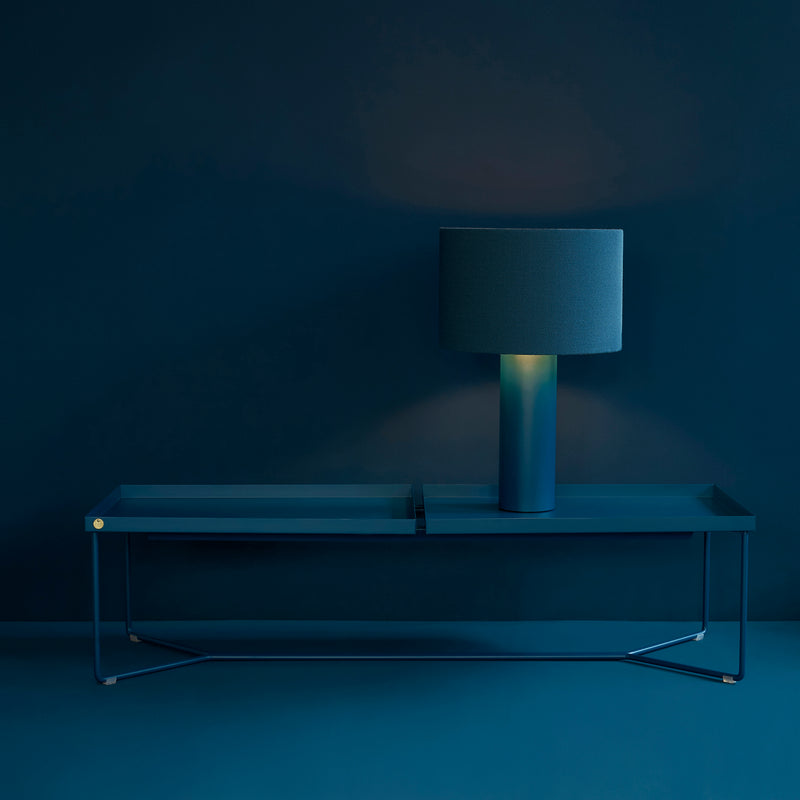 The POI DOUBLE Coffee Table by Victor Foxtrot, in a sleek monochromatic teal design with subtle leather inlays, complements a minimalist lamp with a cylindrical shade. Together against the matching teal wall, they create a serene ambiance with ambient lighting.