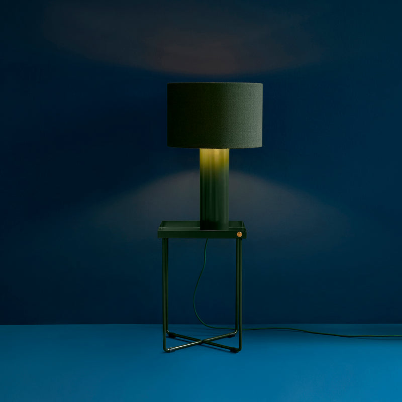 A lit modern black lamp rests on the POI MINI Side Table by Victor Foxtrot, set against a dark blue wall and floor. Its warm glow casts light on the wall, creating a striking contrast with the surrounding dark tones.