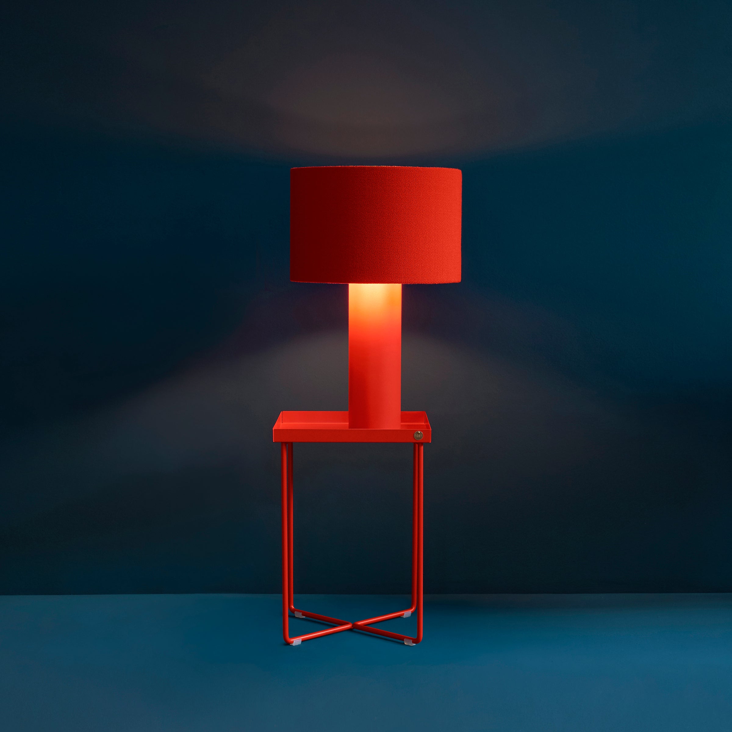 A POI MINI Side Table by Victor Foxtrot, featuring a modern design, showcases a table lamp with a vibrant red lampshade and base. The lamp casts a warm glow against the dark blue background, emphasizing its striking color.