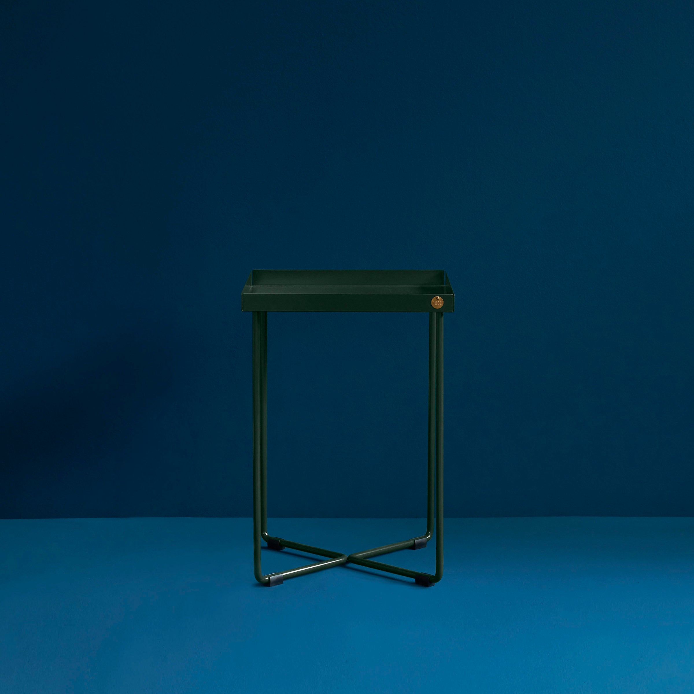 The POI MINI Side Table by Victor Foxtrot features a sleek, modern design with a square green seat and thin metal legs on a cross-shaped base, resembling a stool against a dark blue background while highlighting its elegant, contemporary style.