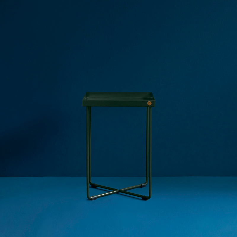 The POI MINI Side Table by Victor Foxtrot features a sleek, modern design with a square green seat and thin metal legs on a cross-shaped base, resembling a stool against a dark blue background while highlighting its elegant, contemporary style.