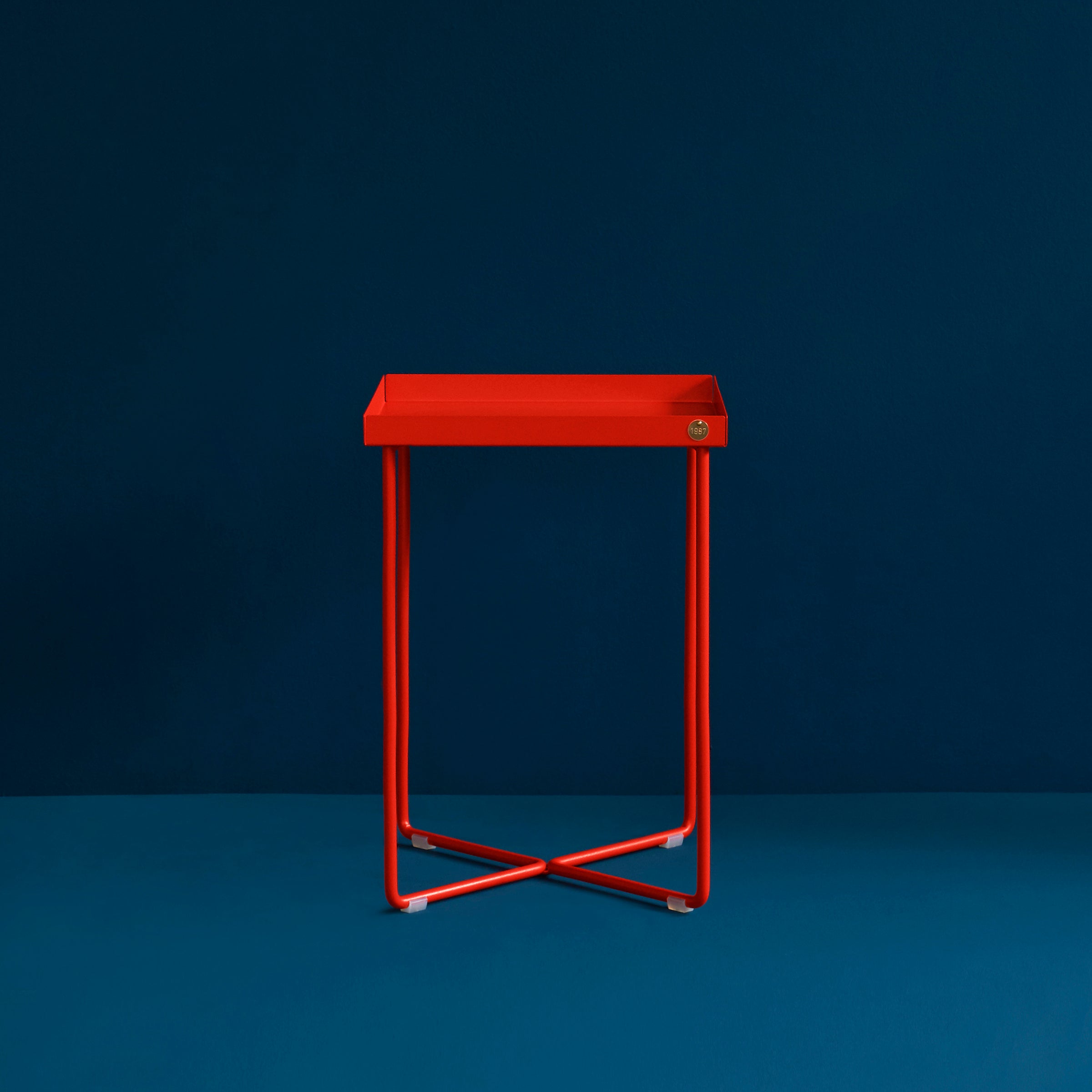 The POI MINI Side Table by Victor Foxtrot is a sleek, minimalist piece. Its slender red legs stand out against a dark teal background, featuring a small emblem for added decorative touch and exuding modern elegance and simplicity.