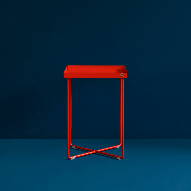 The POI MINI Side Table by Victor Foxtrot is a sleek, minimalist piece. Its slender red legs stand out against a dark teal background, featuring a small emblem for added decorative touch and exuding modern elegance and simplicity.