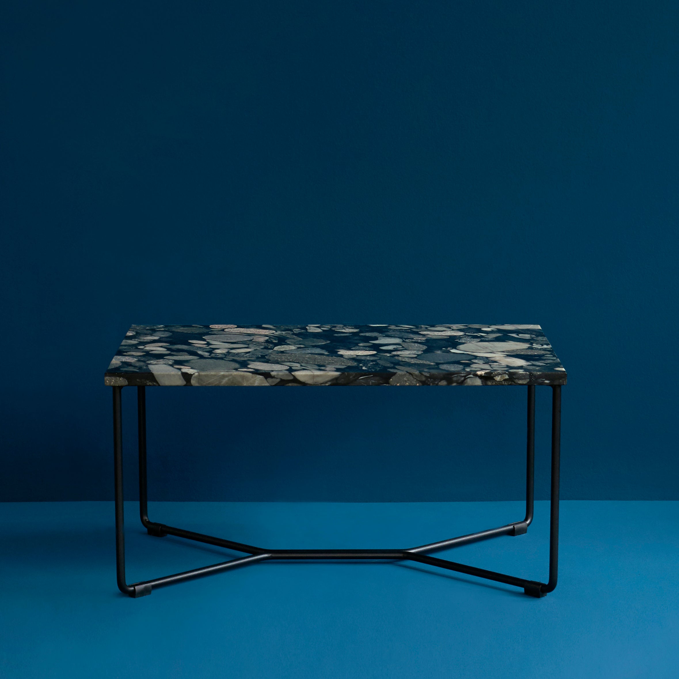 A POI SINGLE MARBLE Side Table by Victor Foxtrot features a rectangular, dark Nero Marinace stone top with unique, irregular spots and thin black metal legs, set against a blue background to enhance its minimalist design.