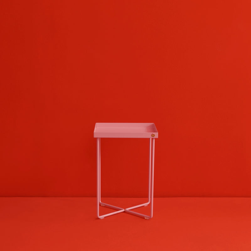 The POI MINI Side Table by Victor Foxtrot, with its minimalist pink square seat and thin metal legs, stands against a vivid red background. Its modern design and small corner accent enhance the striking color contrast, resembling a sleek mini table.