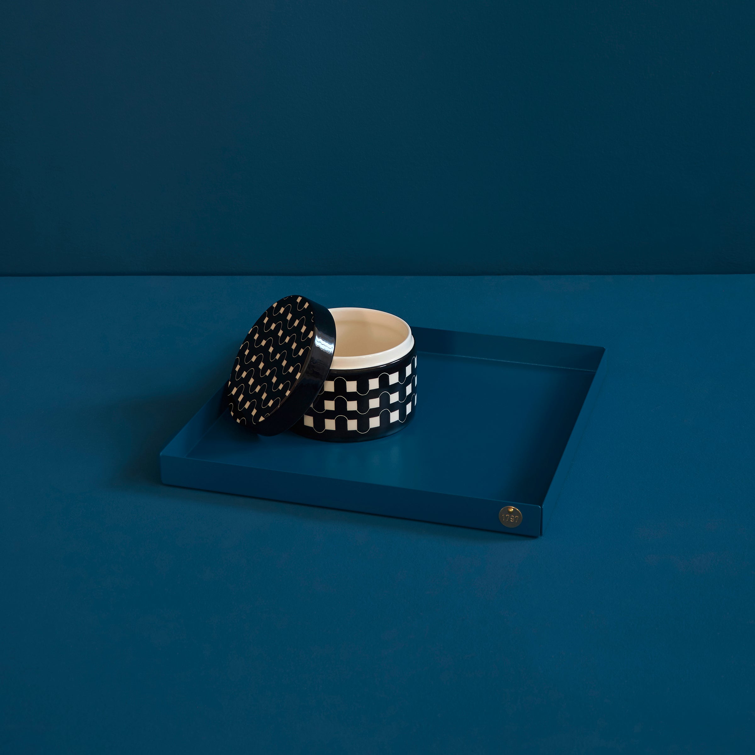 A Victor Foxtrot black and white checkered ceramic container with a geometric lid rests open on a dark blue POI Tray XL, set against a matching background. Ideal for design lovers, the lid is slightly ajar, revealing the cream interior.