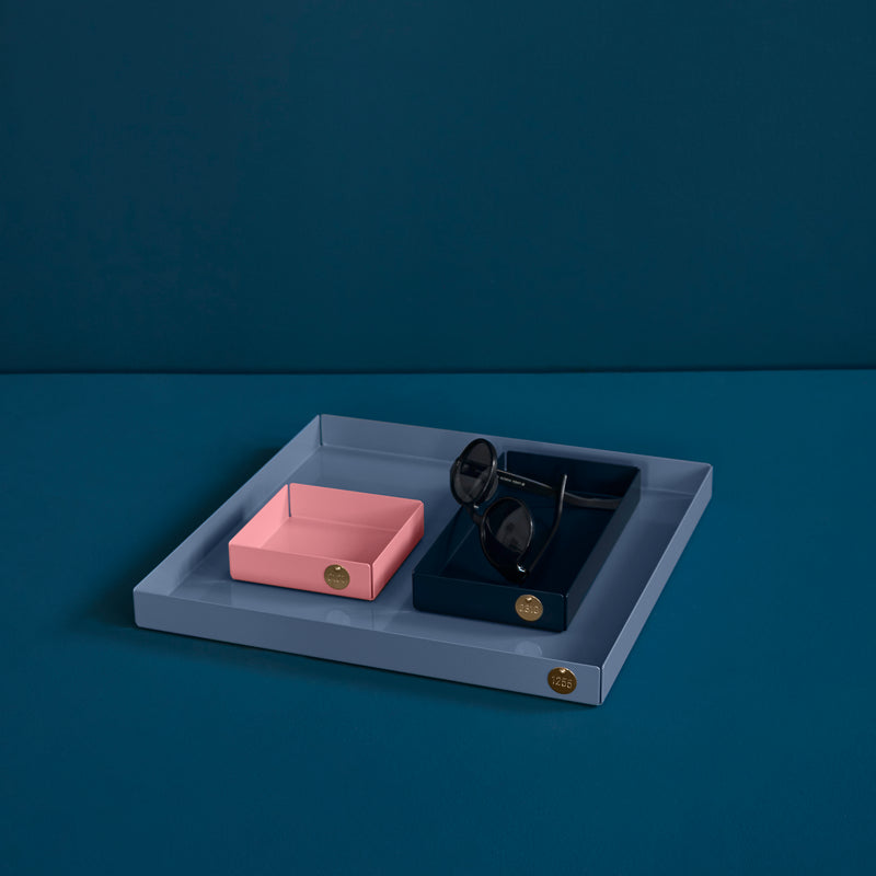Victor Foxtrots POI Tray S is a chic piece for design lovers, featuring a steel blue tray with square pink and dark blue compartments. The dark blue section holds black sunglasses, adorned with gold-colored accents, all set against a deep teal backdrop.