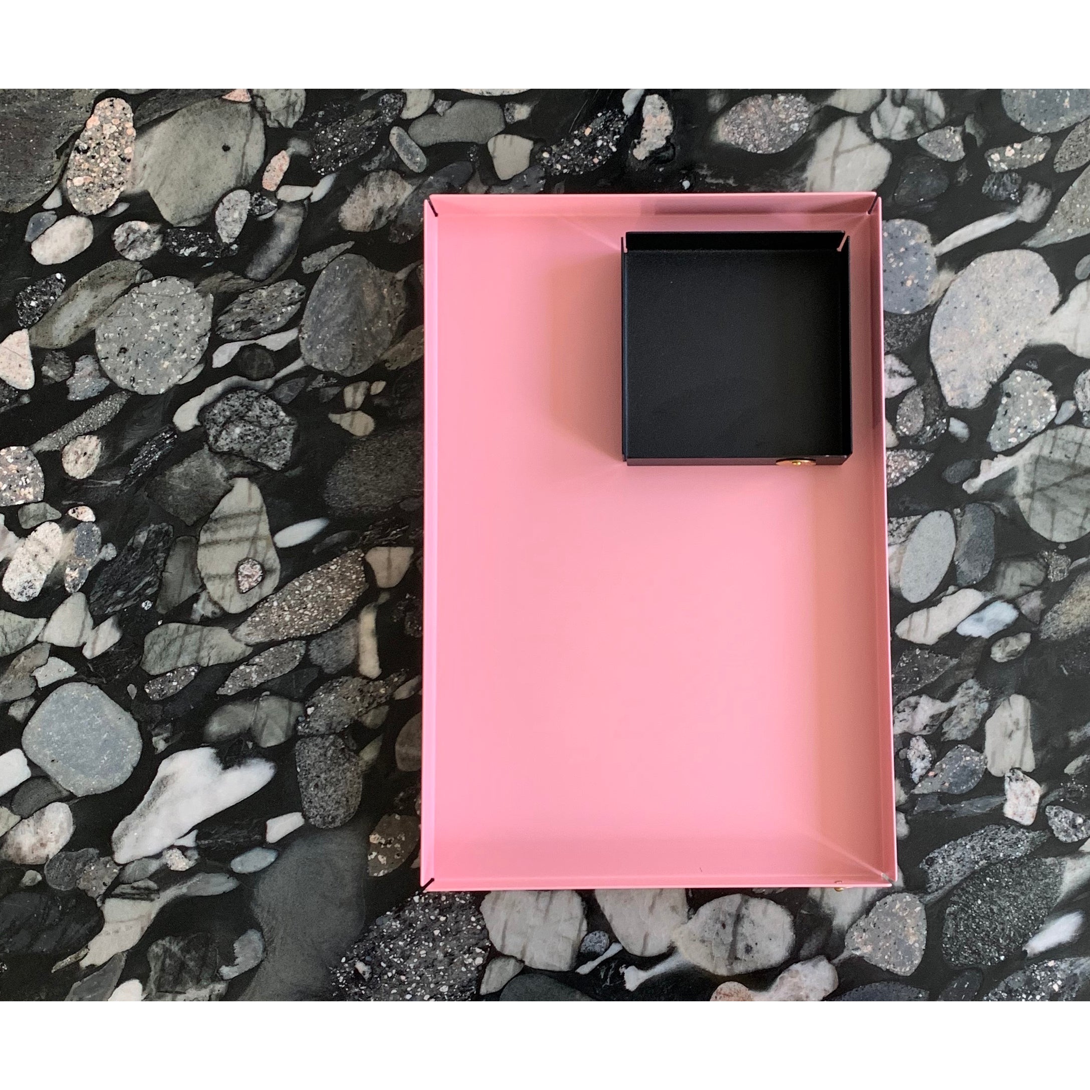 The rectangular pink POI Tray S by Victor Foxtrot, crafted from powder-coated steel, elegantly holds a small square black box on a dark marbled stone surface with intricate grey and white patterns—a must-see for design lovers.
