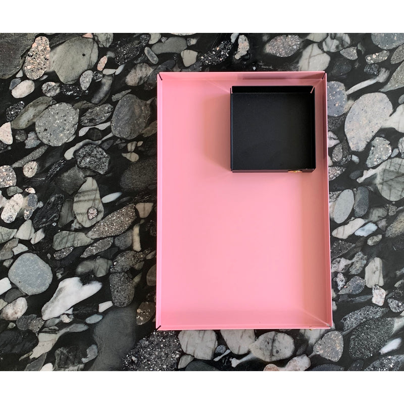 The rectangular pink POI Tray S by Victor Foxtrot, crafted from powder-coated steel, elegantly holds a small square black box on a dark marbled stone surface with intricate grey and white patterns—a must-see for design lovers.