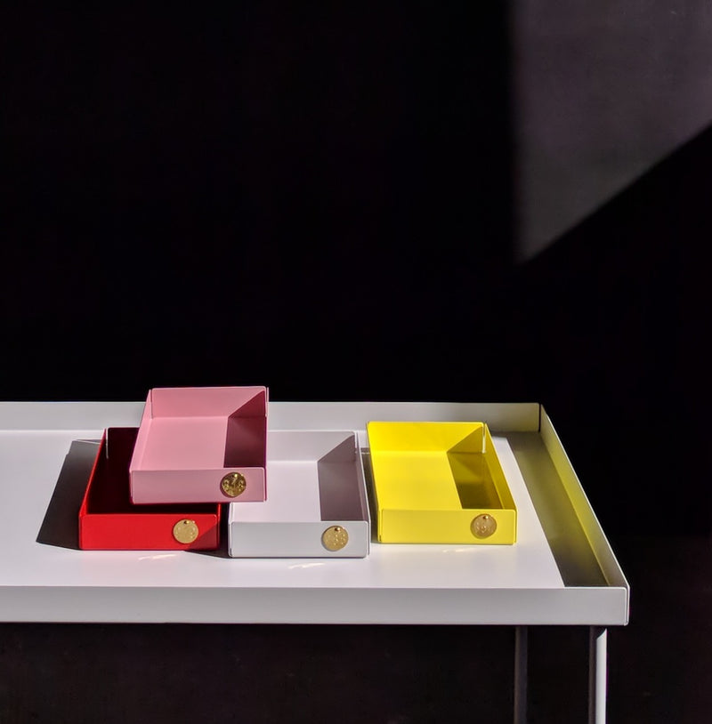 Four vibrant POI Tray M boxes by Victor Foxtrot, crafted from powder-coated steel in red, pink, white, and yellow with gold emblems, are stacked on a white table against a dark backdrop. Shadows create contrast with the bright colors. Ideal for design enthusiasts.