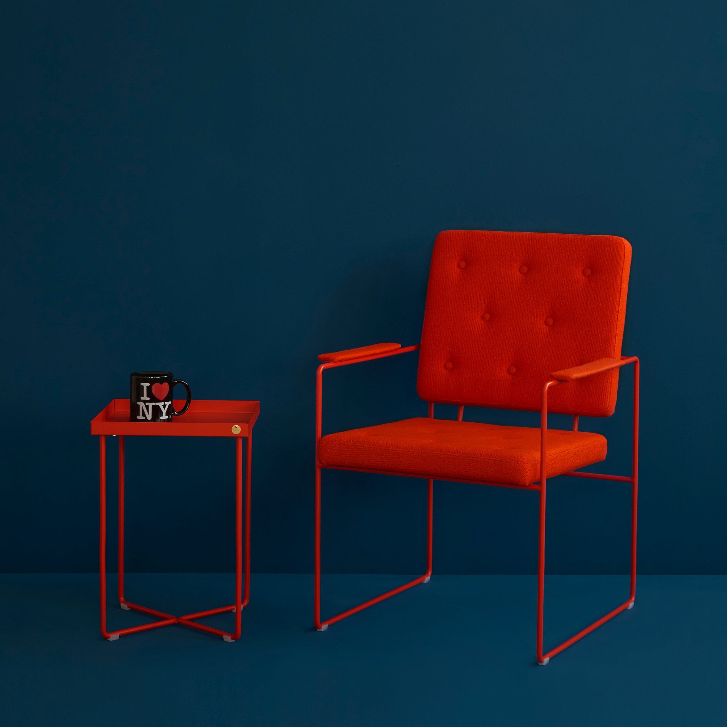 A modern orange armchair with a button-tufted backrest sits next to a Victor Foxtrot POI MINI Side Table. On the table, a black mug with I ♥ NY printed on it complements the deep teal wall in the background.