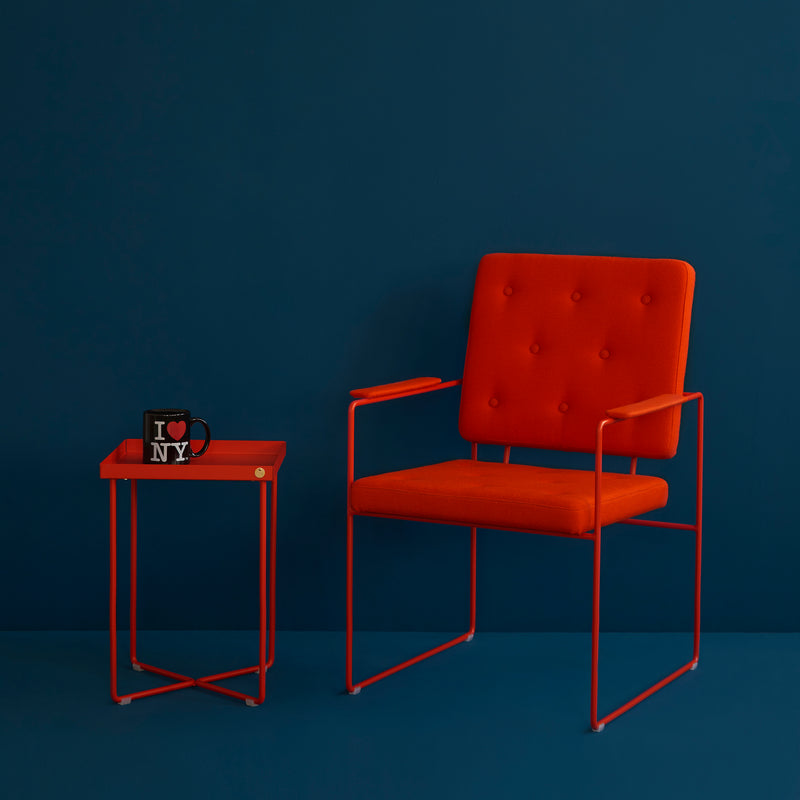 A modern orange armchair with a button-tufted backrest sits next to a Victor Foxtrot POI MINI Side Table. On the table, a black mug with I ♥ NY printed on it complements the deep teal wall in the background.