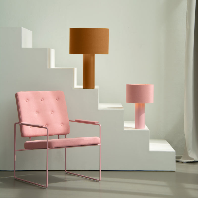 A modern interior scene showcases a pink armchair with metal legs near white platforms. Each step displays a stylish table lamp—one large with a wool fabric mustard lampshade, and the smaller, pink ALL ROUND MINI by Victor Foxtrot. Sunlight softly illuminates the setting.