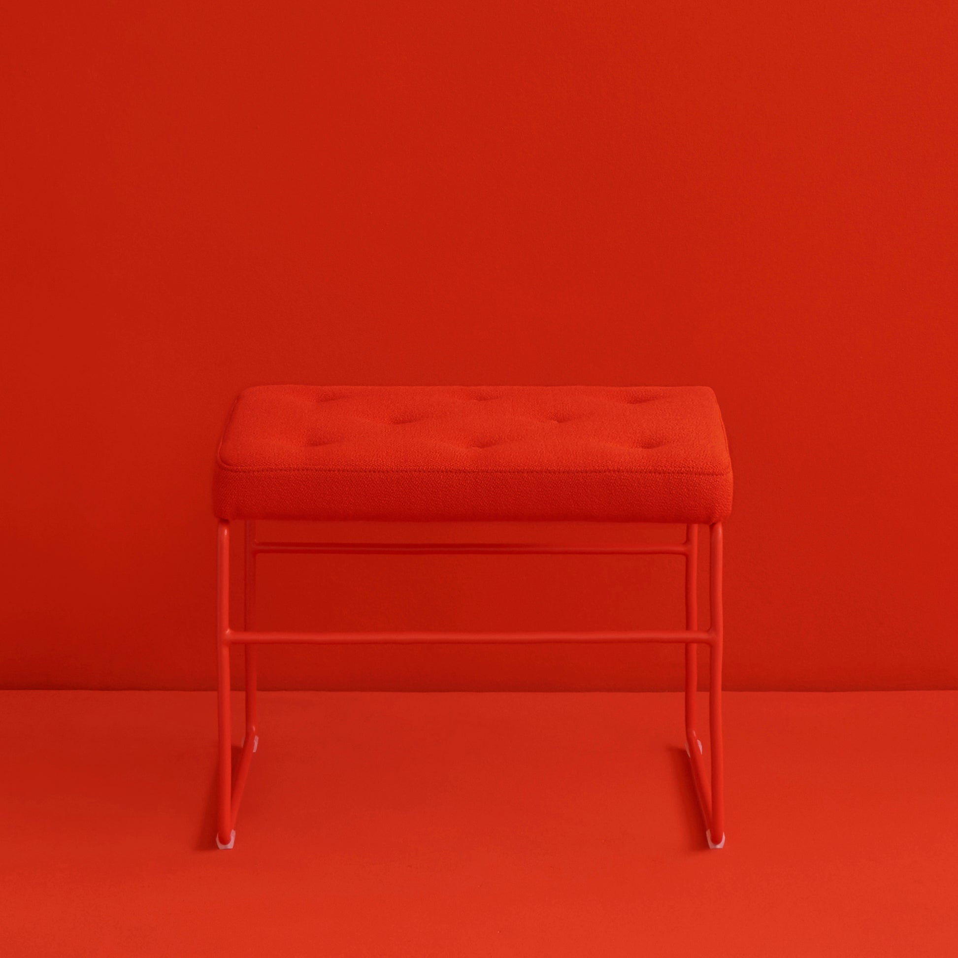 The SWELL TIME Ottoman by Victor Foxtrot features a red rectangular tufted cushion and button stitching on a sleek steel frame with sled legs, resting against a matching background and floor, reflecting the elegance of British upholstered furniture.