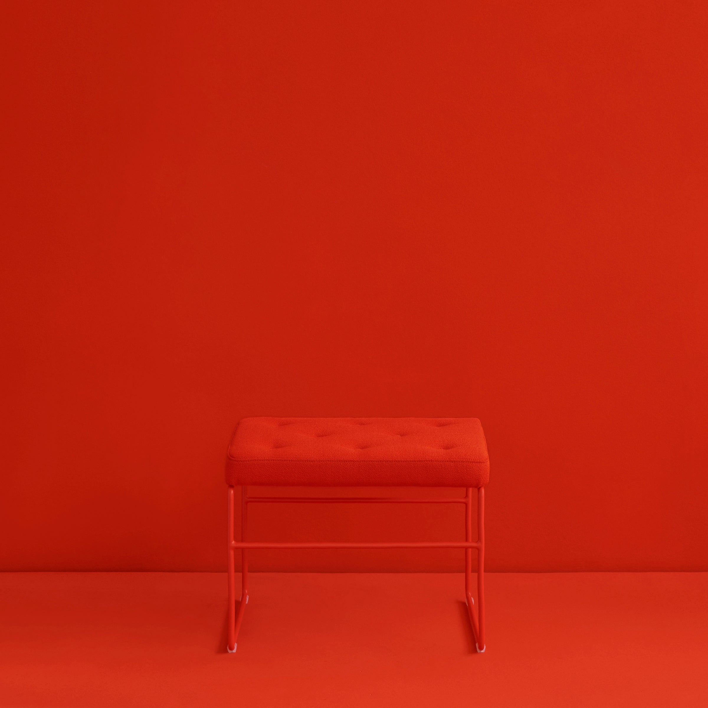 Against a red background, the SWELL TIME Ottoman by Victor Foxtrot creates a monochromatic effect with its small, rectangular design and steel frame. The ottoman showcases button stitching on its tufted cushion top.