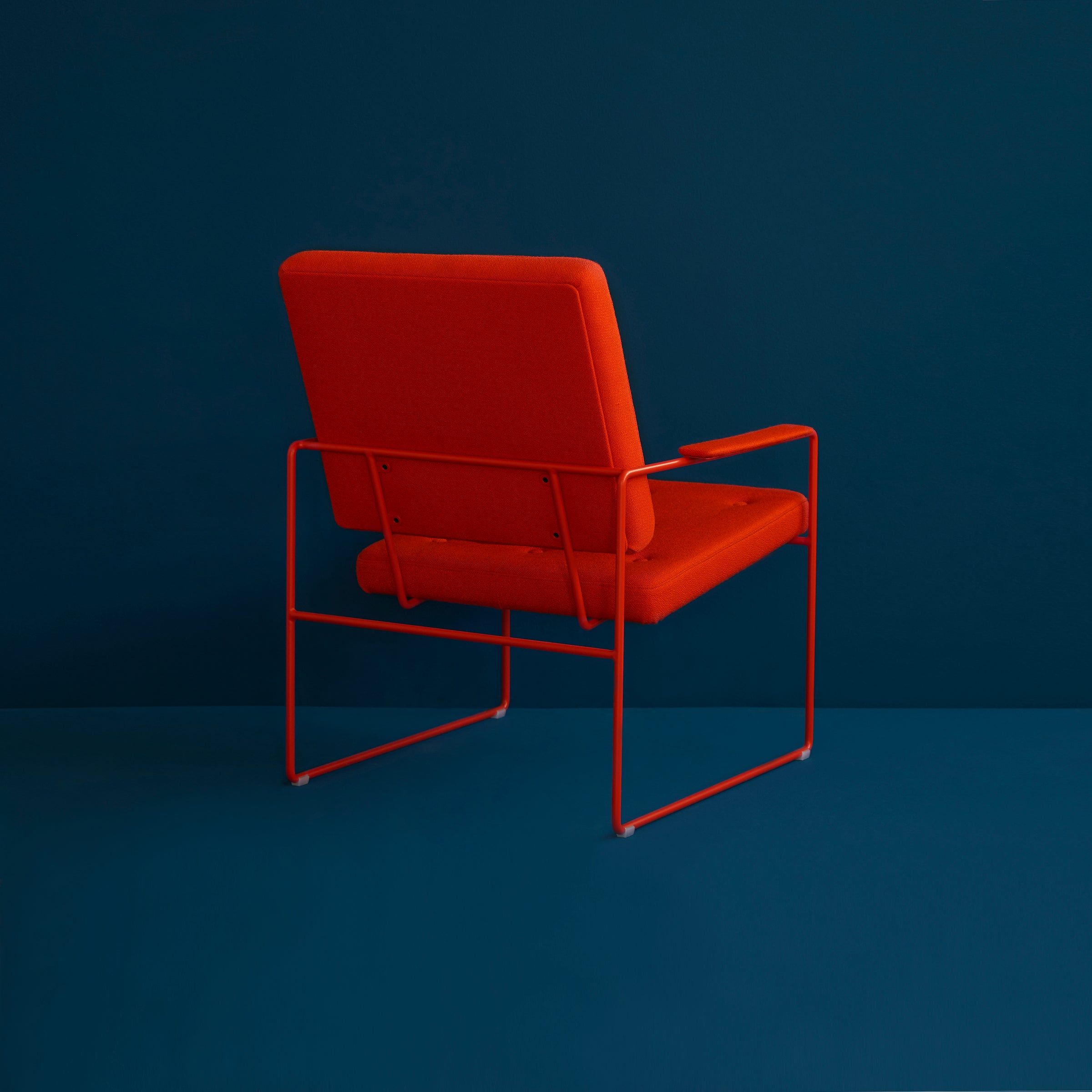 The SWELL TIME Lounge Chair by Victor Foxtrot is a modern designer chair with an orange cushioned seat and backrest, featuring a sleek steel frame. Its positioned facing away from the viewer against a solid dark teal background.