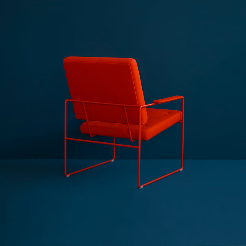 The SWELL TIME Lounge Chair by Victor Foxtrot is a modern designer chair with an orange cushioned seat and backrest, featuring a sleek steel frame. Its positioned facing away from the viewer against a solid dark teal background.