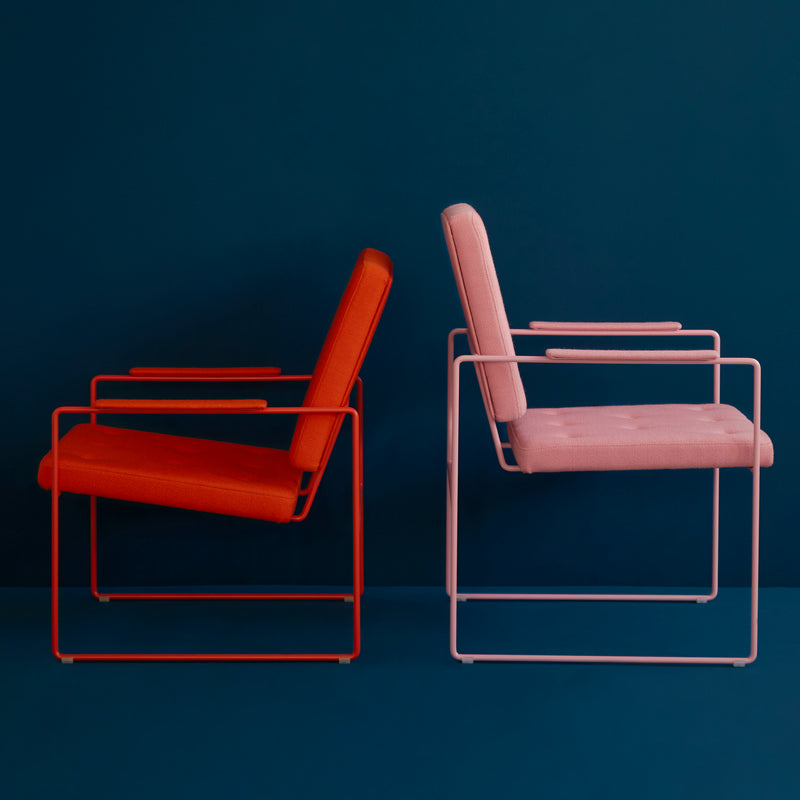 Two SWELL TIME Lounge Chairs from Victor Foxtrot, featuring sleek steel frames and expert British upholstery, are positioned against a dark teal backdrop. The left chair has a vibrant red cushion, while the right boasts a soft pink one, highlighting their minimalist design.