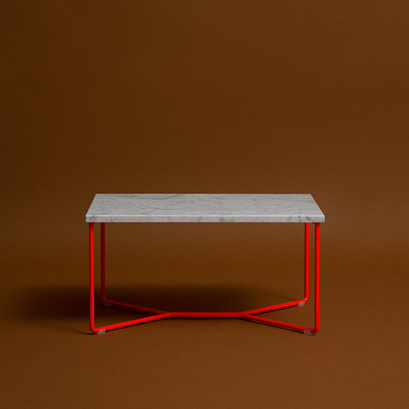 The POI SINGLE MARBLE Side Table by Victor Foxtrot features a sleek rectangular white marble top with a striking red steel frame, set against a warm brown backdrop.
