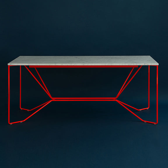 The SAME SAME Marble Table by Victor Foxtrot showcases a minimalist design with a smooth, light surface akin to Carrara Venatino marble. Set against a dark background, its geometric red powder-coated steel frame offers a striking contrast.