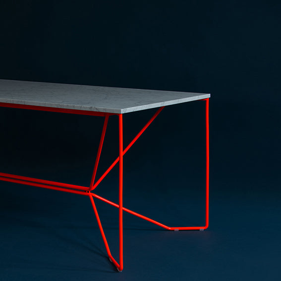 The Victor Foxtrot SAME SAME Marble Table showcases a minimalist design with a light gray rectangular tabletop resembling Carrara Venatino marble and bright red powder-coated steel geometric legs set against a dark background.