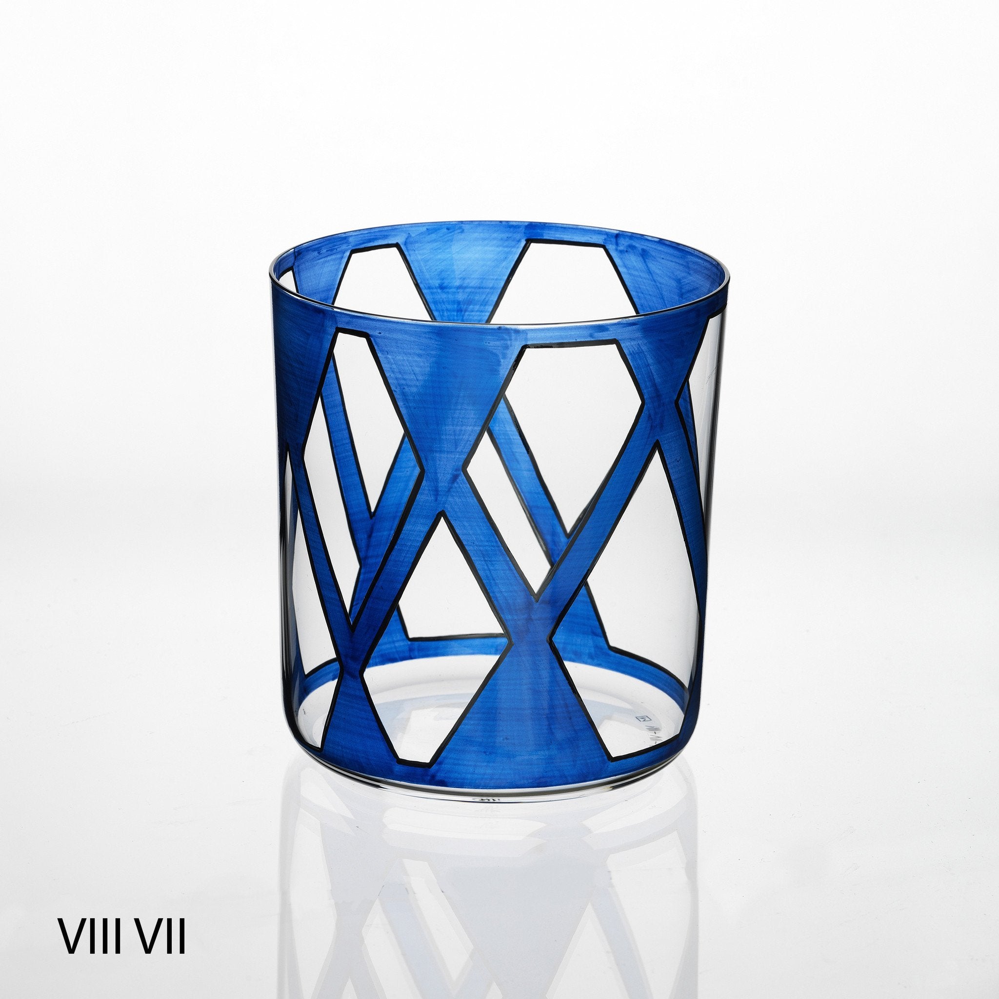 The Neo Light VIII Enamel Collection by Lobmeyr features a transparent cylindrical glass with blue geometric interlocking diamond patterns, reflecting Martino Gampers contemporary ornament style. It displays VIII VII in Roman numerals and stands elegantly on a reflective white surface.