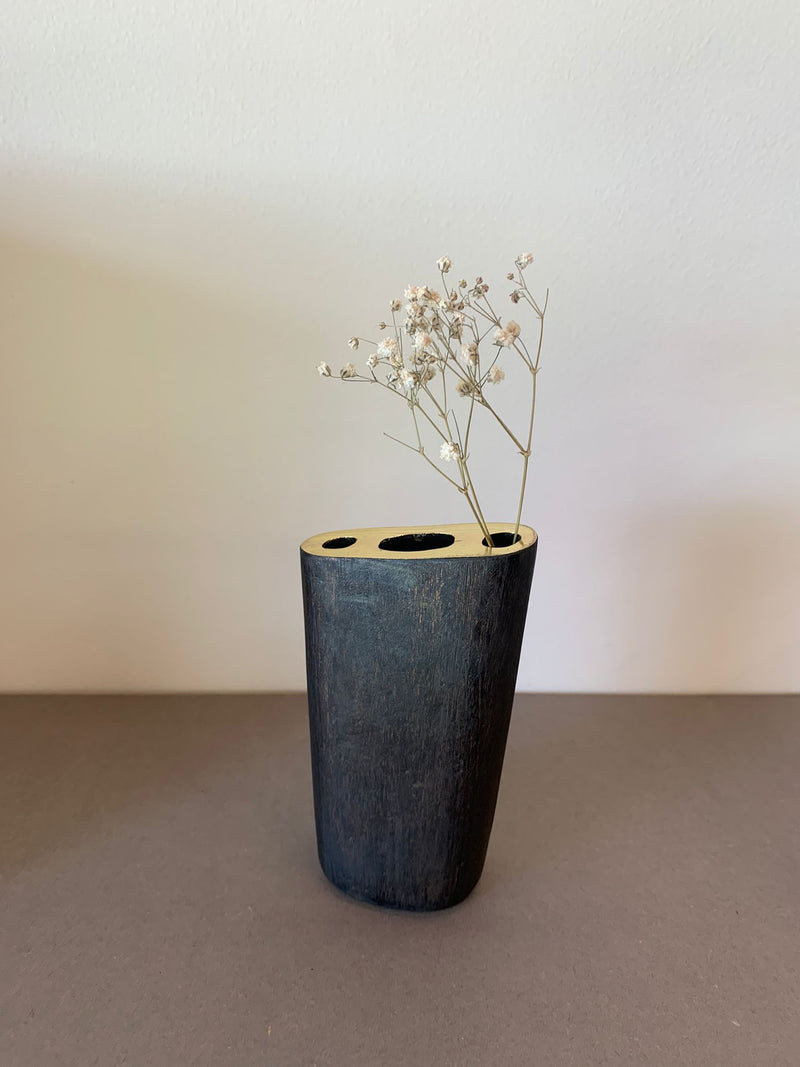 A minimalist black Vase #7235 - Sale by Werkstätte Carl Auböck, featuring a smooth matte finish, holds small white flowers against a plain background, evoking simplicity and elegance.