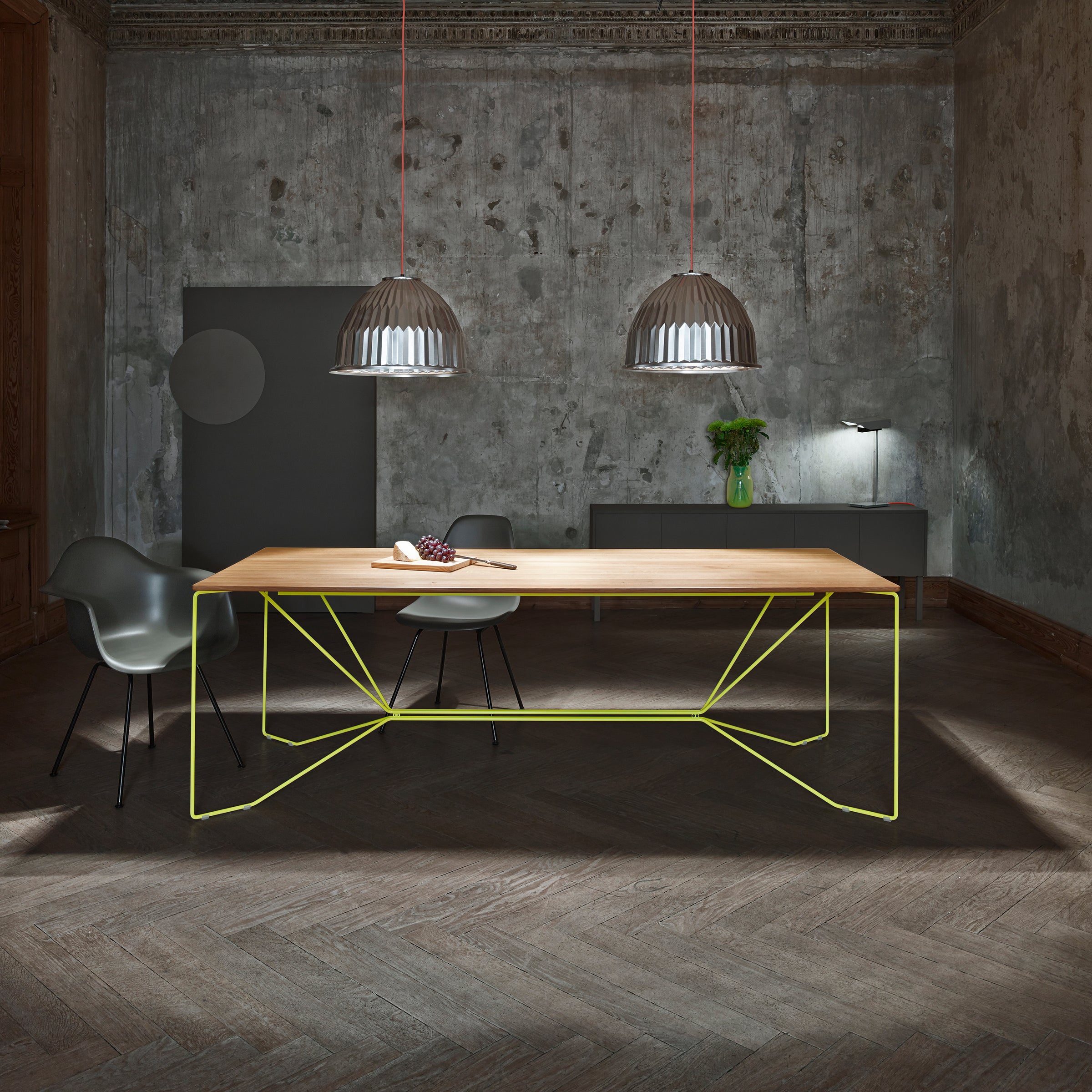 In a modern dining room, the SAME SAME Oak Table by Victor Foxtrot features artistic yellow powder-coated steel legs. Two pendant lights illuminate gray walls and a sideboard with a vase of greenery, while two dark chairs complete the chic minimalist decor.